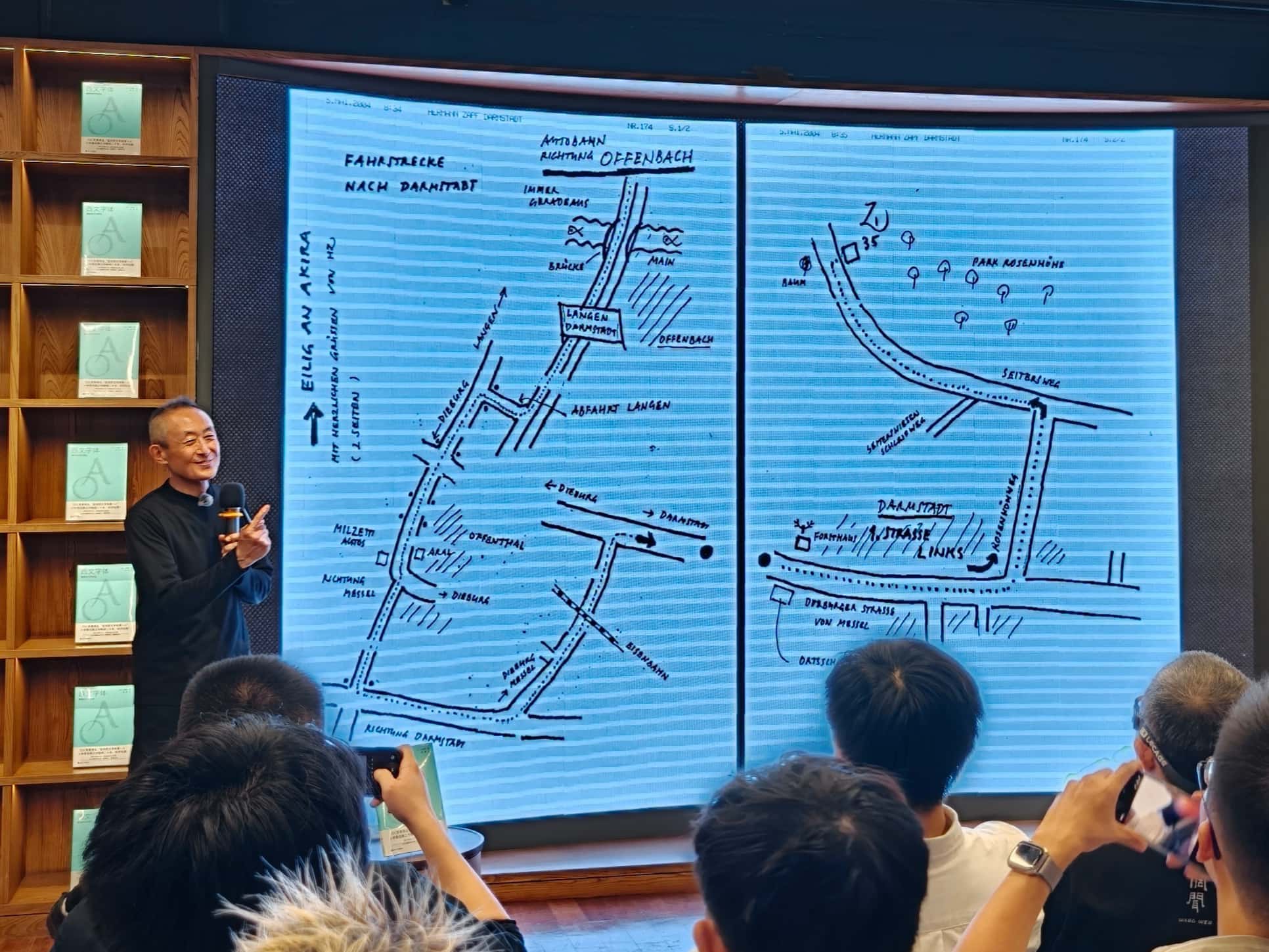 小林章在台上展示两页手绘路线图,台下观众举起手机拍摄。
