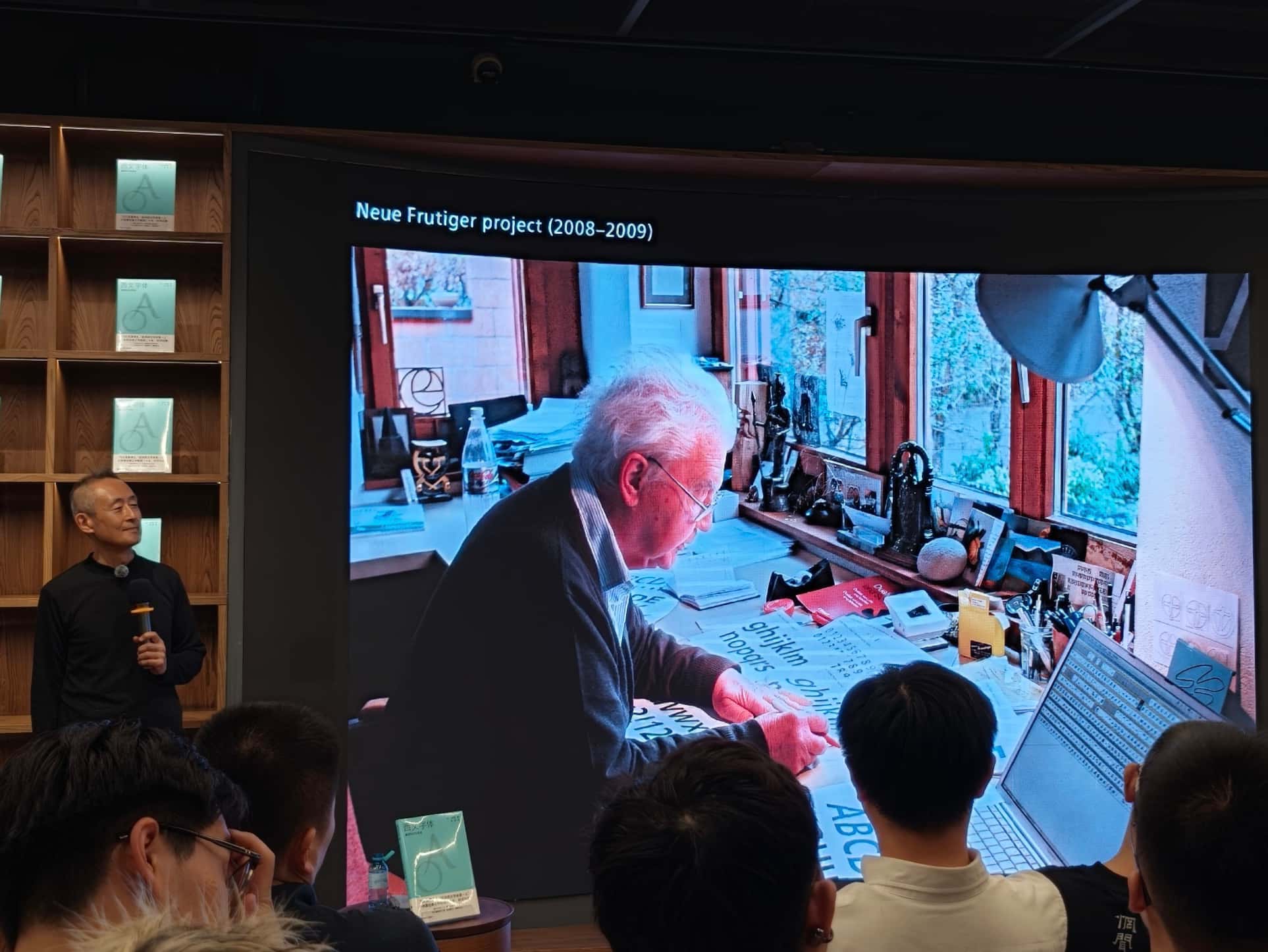 小林章站在台侧,投影幕显示弗鲁提格伏案绘制 Neue Frutiger 字样。